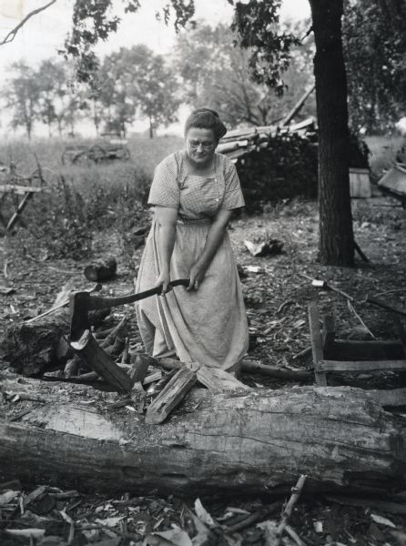 A woman using an axe to chop wood in a rural area. A pile of logs is in the background.