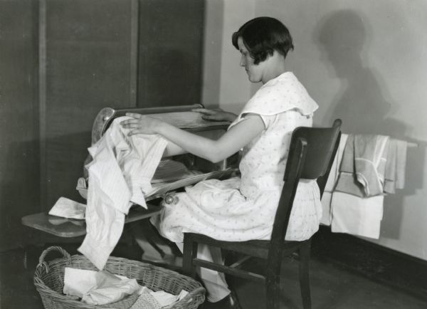 A woman sitting in a wooden chair while using an electric iron on clothing. A laundry basket is at her feet.