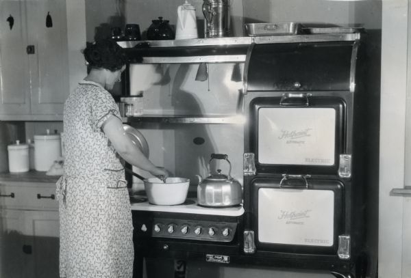 A woman stirs a pot on a six-burner electric stove. Two Hotpoint automatic electric ovens stand beside the stove, and canisters and teapots stand on the stove and on surrounding counters and shelves.