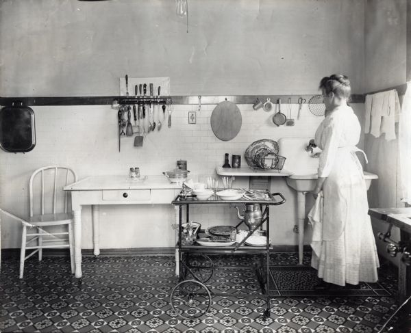 A woman wearing an apron standing near a wheeled butler's tray in a kitchen. A sink, table, and chair are standing against the wall, and various cooking utensils and dishes are on the table and on wall hooks.