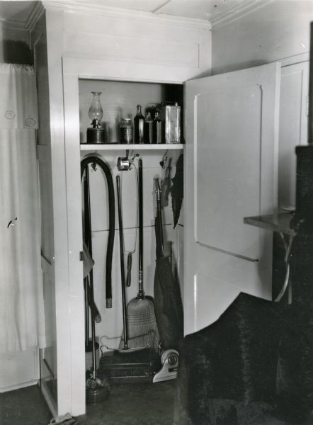 View into a storage closet in the kitchen of Marilla Zearing. A vacuum cleaner, broom ("bissel"), kerosene lantern, and other supplies are stored inside, and a pencil sharpener is attached to the closet shelf.