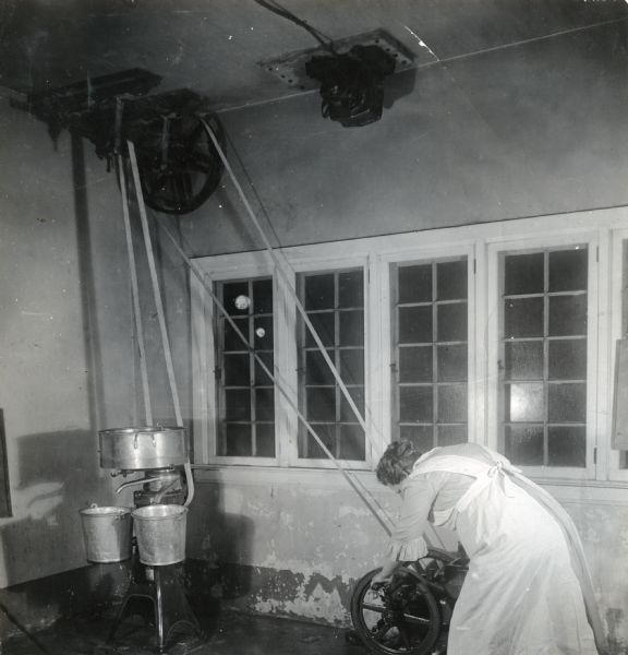 Mrs. Robert Viall is using a gasoline engine to run a cream separator on the Babcock farm. The cream separator is driven by belts that are attached to the engine by pulleys attached to the ceiling.