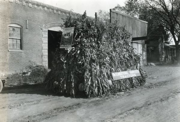 A parade float built on a Farmall tractor parked outside a building on a dirt road. The tractor is covered in corn stalks and has signs on the front and side that read: "McCormick-Deering Farmall. A Farmall on Every Farm" and "International Harvester Tractors."