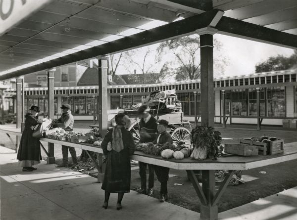 A man and two boys are standing behind an outdoor counter while selling farm produce to female customers. A horse-drawn wagon is parked behind them.