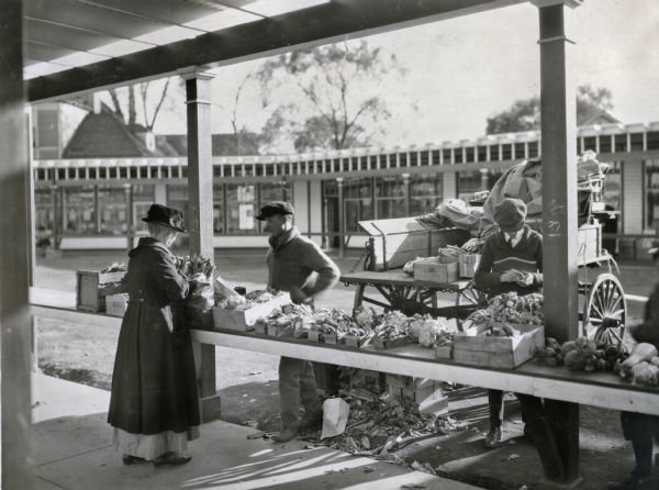 Various types of vegetables are arranged along a counter in an outdoor marketplace. A female customer purchases a bag of farm produce from a male vendor; another man stands nearby, and a horse-drawn wagon loaded with additional merchandise is parked behind them.