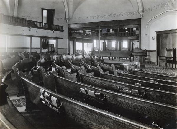 Interior view looking toward the altar across rows of pews inside the Church of Brethren in Orange Township, Black Hawk County. The church appears to consist of several wings, including separate rooms with curtains pulled back. A balcony curves around the upper level of the building.
