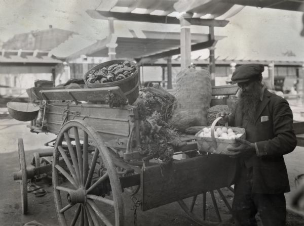 A man, probably a farm vendor, standing next to a horse-drawn wagon loaded with farm produce at what appears to be an outdoor marketplace.  The man is wearing a derby and is holding a basket of chicken eggs.