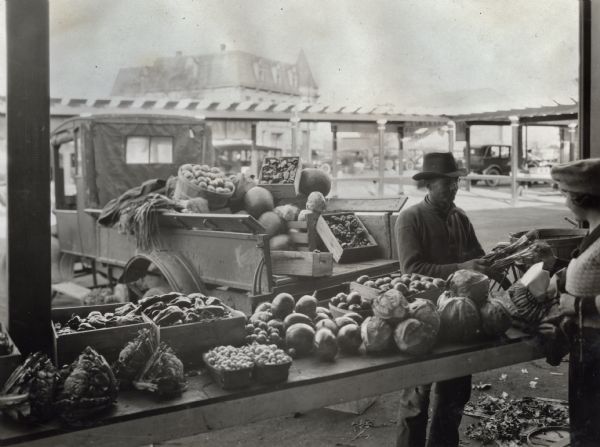 A man selling green onions to a female customer from behind a produce-laden counter at an outdoor market. A truck loaded with additional merchandise is parked behind him. A multi-story building and other vendor stalls are in the background.