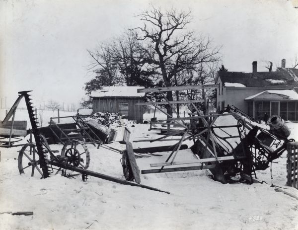 Farm equipment, including a grain binder and mower, are lying in the snow outside a farmhouse. In the background on the right two children are standing near the screened porch of the house. A shed and log pile are on the left.