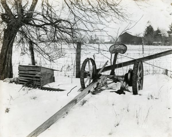 A horse-drawn mower lying in snow near a wire fence. A barn and several other farm buildings are in the background beyond a field. A wooden crate stands beneath a tree beside the mower.