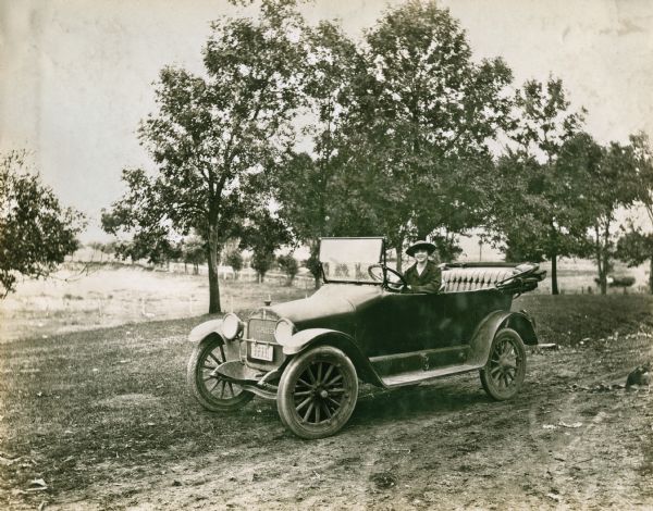 A woman wearing a wide-brimmed hat sits behind the wheel of a Case automobile with its top folded down. A chicken or rooster stands to the right along the side of the rural dirt road.