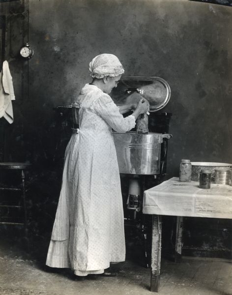 A woman wearing a dress and bonnet is using a wire potato masher to remove a jar of peaches from a vat of boiling water. Several jars of canned goods are standing on the table covered in newspaper beside her, and a wooden stool is standing to her left.