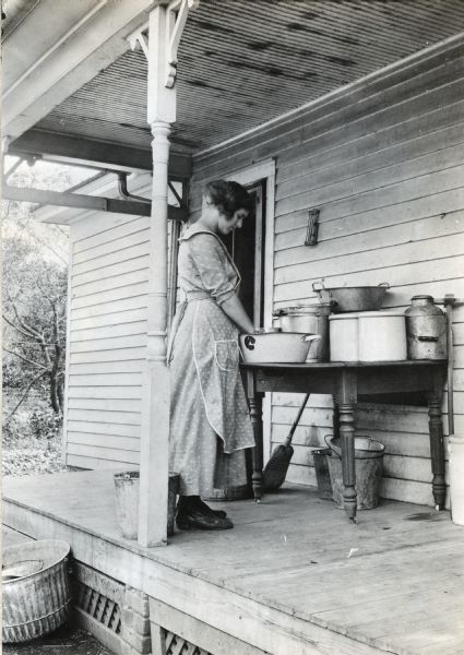 A woman wearing a dress and apron washes her hands in a metal bucket set on a table on a farmhouse porch. Multiple metal pails with handles and a milk pail are stacked on the table, and a broom rests against its side.