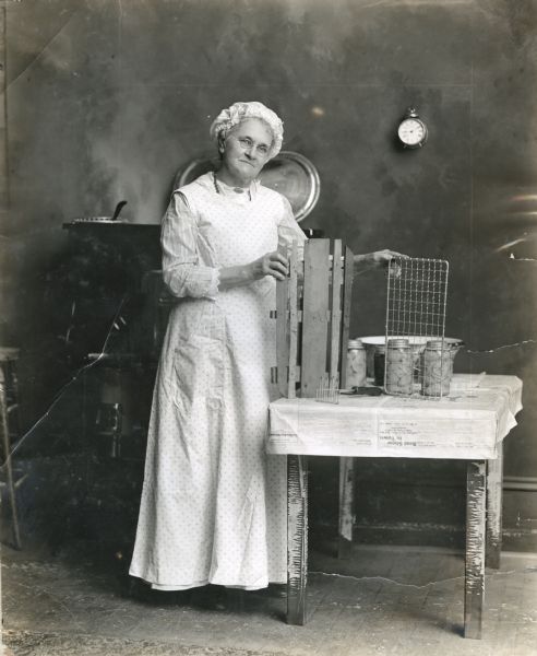 Mrs. Buckley, wearing a dress, apron, and bonnet, is standing next to a table where equipment used for the home canning of agricultural produce is displayed on a table covered in newspaper. She is holding two racks for use in the boiler that is standing behind her, one wooden and one metal.