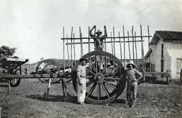 Two men standing by the wheel of a wooden horse-drawn wagon while another man is standing behind wooden bars on the wagon bed. A barn, additional buildings, and other farm equipment are in the background.