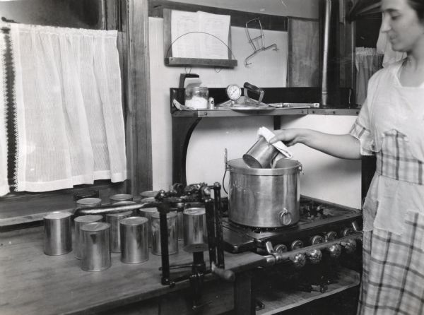 A woman is using a towel to remove a can from a stove top pressure boiler.  Additional cans are sitting on the counter beside her, and the boiler lid and other tools are on a shelf above the stove.