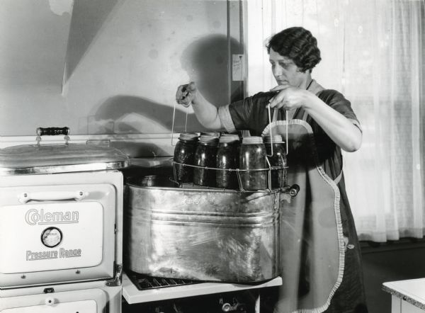 A woman wearing an apron is removing a wire rack filled with glass canning jars from a boiler on a Coleman Pressure Range on International Harvester's Hinsdale experimental farm.