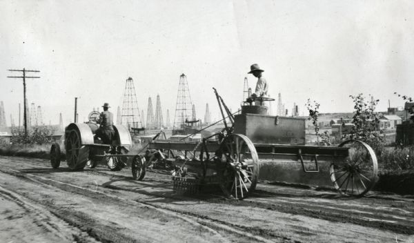 Two men use a 15-30(?) tractor and a grader on a dirt road. In the background are numerous oil derricks and industrial buildings.