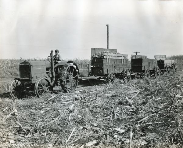 Delmar Van Horn uses a McCormick-Deering tractor to pull three wagons full of corn across a cut field.