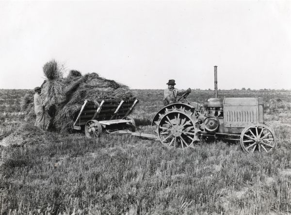 A man piles hay on a wagon pulled by a 10-20(?) tractor driven by a man sitting behind the wheel. The photograph was taken on International Harvester's Hinsdale experimental farm.