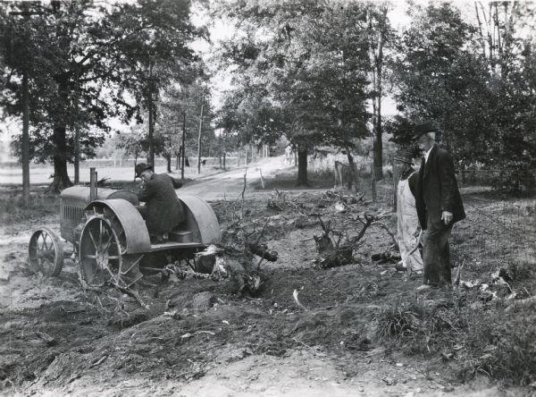 A man is using a 10-20(?) tractor and chain to remove a stump from a piece of land on the R.R. Robertson farm. Two other men are looking on.