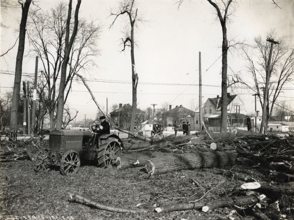 A man is using a McCormick-Deering 10-20(?) tractor to assist in clearing land in a residential area. Several other men are standing in the background holding axes.