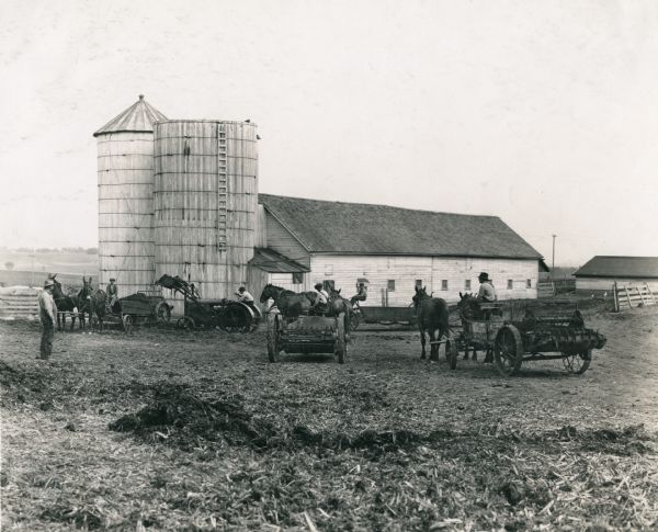 A group of men use a tractor and horse-drawn manure spreaders in a barnyard on the farm of J.C. Witmer. The tractor appears to have a "Lessman" loader.