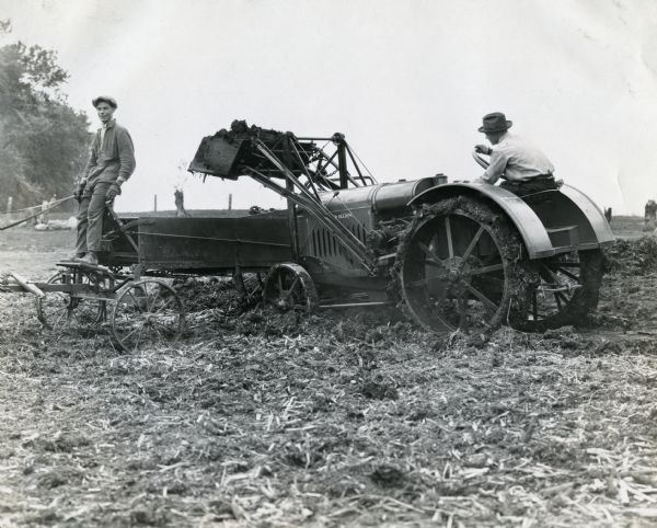 A man using a McCormick-Deering 10-20 tractor outfitted with a Lessman loader to load a manure spreader. Another man is standing on the front of the loader.