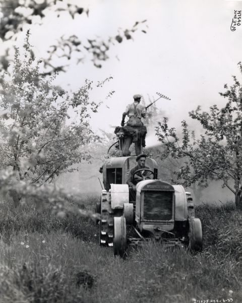 A man smoking a cigarette drives a McCormick-Deering 10-20 tractor as another man stands on the tank of an attached 150-gallon sprayer while applying pesticide to orchard trees. The photograph was taken in Markham, Ontario, Canada.