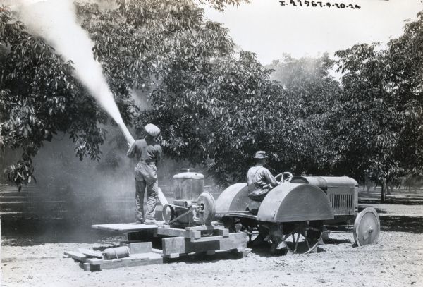 A man stands on a platform made of wood while using a sprayer to apply pesticide to a grove of fruit trees. Another man stands on the back, and another man is driving. The sprayer is powered by a McCormick-Deering 10-20 orchard tractor.