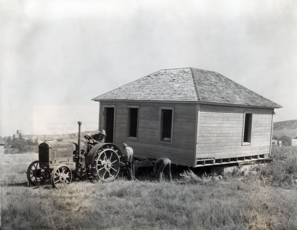 Three men use a McCormick-Deering 15-30(?) tractor to move a wooden building, possibly a small home or shed, with a trailer.