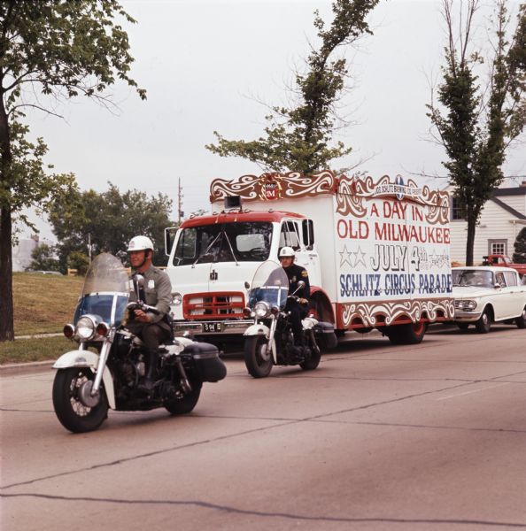 A calliope truck with text reading "Jos. Schlitz Brewing Co. Presents / A Day in Old Milwaukee / July 4th / Featuring Schlitz Circus Parade" drives down a road in a residential area accompanied by two police escorts on motorcycles. The truck was part of a 25-piece circus parade that traveled from Circus World Museum in Baraboo to Milwaukee.