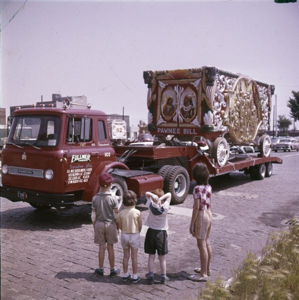 Four children are standing looking at a circus wagon labeled "Pawnee Bill" which is chained to the bed of an International truck. The wagon was part of a 25-piece circus parade that traveled from Circus World Museum in Baraboo to Milwaukee.