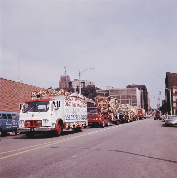 An International truck marked "A Day in Old Milwaukee. July 4th Schlitz Circus Parade" leads a fleet of circus vehicles through a downtown area. The 25-piece parade traveled from Circus World Museum in Baraboo to Milwaukee.