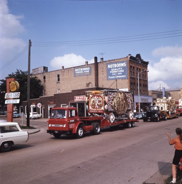 The "Pawnee Bill" circus wagon passes Notbohms furniture store and funeral home along a commercial street as a child looks on from the foreground. Multiple other circus wagons are traveling down the road, carried on the beds of International trucks as part of a 25-piece circus parade from Circus World Museum in Baraboo to Milwaukee.