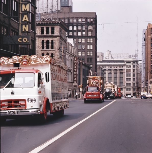 An International CO-192 truck leads a 25-piece circus parade through downtown streets. The parade originated at Circus World Museum in Baraboo. Additional circus wagons are carried on beds of trucks past storefronts advertising sportswear and luggage. Gimbels Department Store stands in the background.