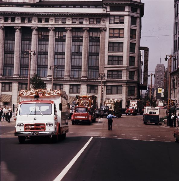 A police officer directs a parade of circus wagons carried on International trucks through downtown streets. The 25-piece parade originated at Circus World Museum in Baraboo. Gimbels Department Store stands in the background.
