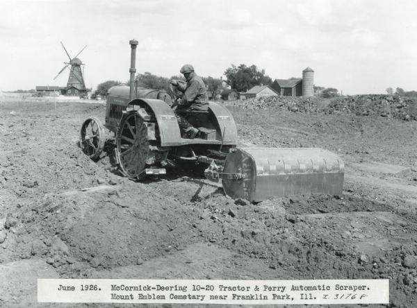 A man uses a McCormick-Deering 10-20 tractor and a Perry automatic scraper to prepare the grounds at Mount Emblem Cemetery. Farm buildings are  in the background, including a large windmill.