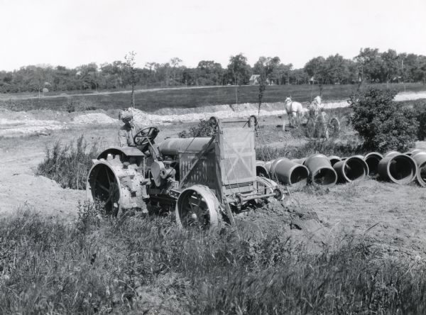 A man uses a McCormick-Deering 10-20 tractor and a Monarch loader to do construction work, possibly irrigation, in a field. Pieces of pipe lie on the ground beside him and another man uses a horse-drawn wagon in the background.