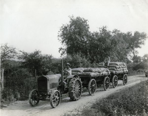 A man uses a McCormick-Deering 10-20(?) tractor to haul two wagons loaded with burlap bags of farm produce, possibly oranges. The tractor drives along a dirt road, passing by what appears to be an orchard; another man rides on one of the wagons.