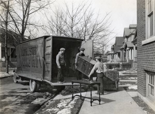 Men remove furniture from a Model 53 International motor truck owned by the Northwestern Moving Company of Detroit, Michigan. An end table and upholstered chair sit on the sidewalk alongside the truck, and residences line both sides of the street.