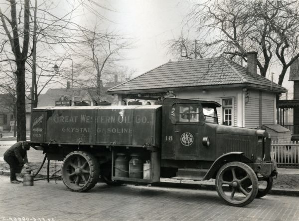 A Model 103 International truck owned by the Great Western Oil Company of Cleveland, Ohio parked on a cobblestone street in a residential area. A man stands at the back of the truck, filling a metal pail with fuel. Behind the truck is a small building with a sign for the Great Western Oil Company hanging under a tiled roof.