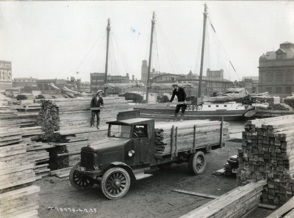 Elevated view of men from the Western Maryland Lumber Company loading lumber onto the back of a Model 63 International truck. Piles of lumber are arranged throughout the lumberyard and several warehouses and industrial buildings are in the background. Ships are in a canal or river behind the lumberyard.