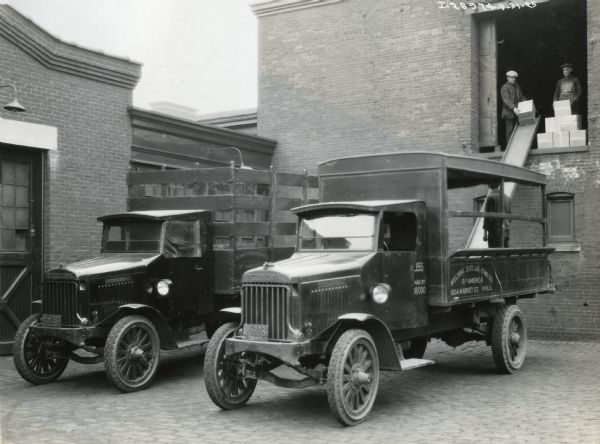 Two men use a slide to load boxes from the loading dock on the second floor of the Welsbach Street Lighting Company onto the bed of one of the company's Model 63 International trucks. Another truck is parked alongside it in the loading dock area.