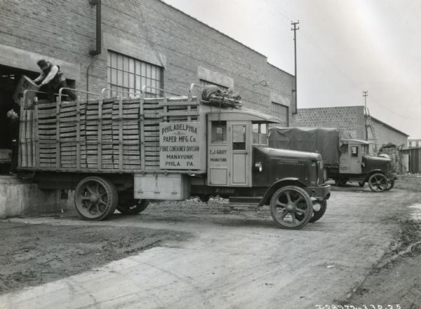 Men remove stacks of cardboard from the back of a Model 103 International truck at the loading dock of E.J. Graff. Another truck is parked in the background.