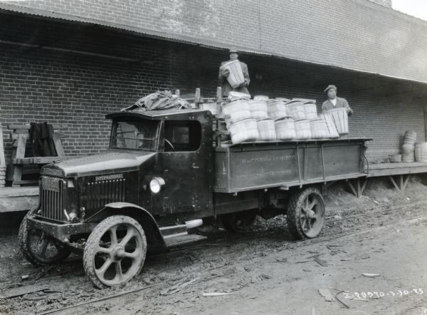 Men loading bushels of fruit and vegetables onto the back of a Model 63 International truck owned by the McCray & Hunter company. A loading dock of a brick building is behind the truck.