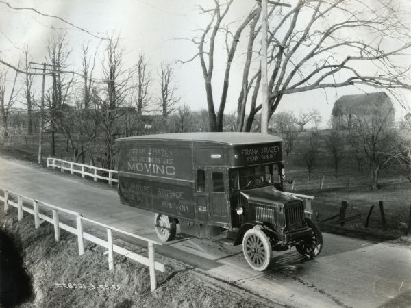 Elevated view of a Model 63 International moving van owned by Frank Razey Moving Company driving down a rural highway. A barn is in the background.