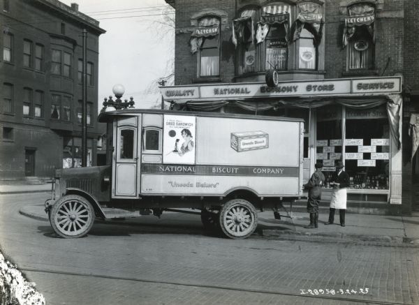 Two men stand at the rear of a Model 43 International truck owned by William Millard and equipped with a National Biscuit Company body. The truck is parked in front of Fletcher's National Economy Store.