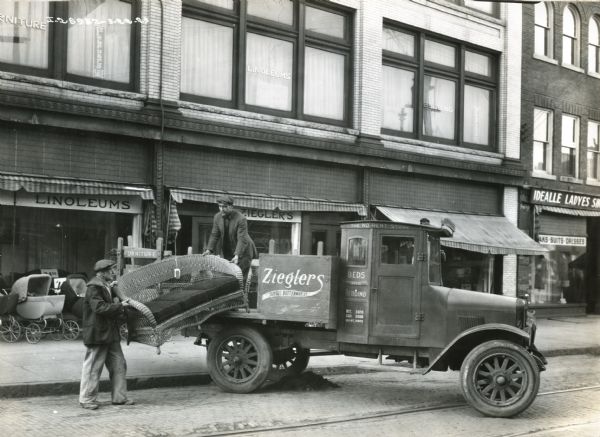Two men are unloading a wicker sofa from the back of a Model S International truck owned by Zieglers Furniture Store.