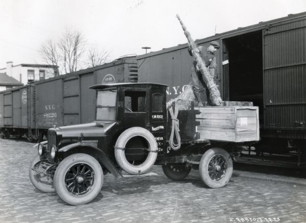 An International Model S truck owned by T.W. Rice is parked alongside an opened railroad boxcar as a man unloads a plant from the truck bed.